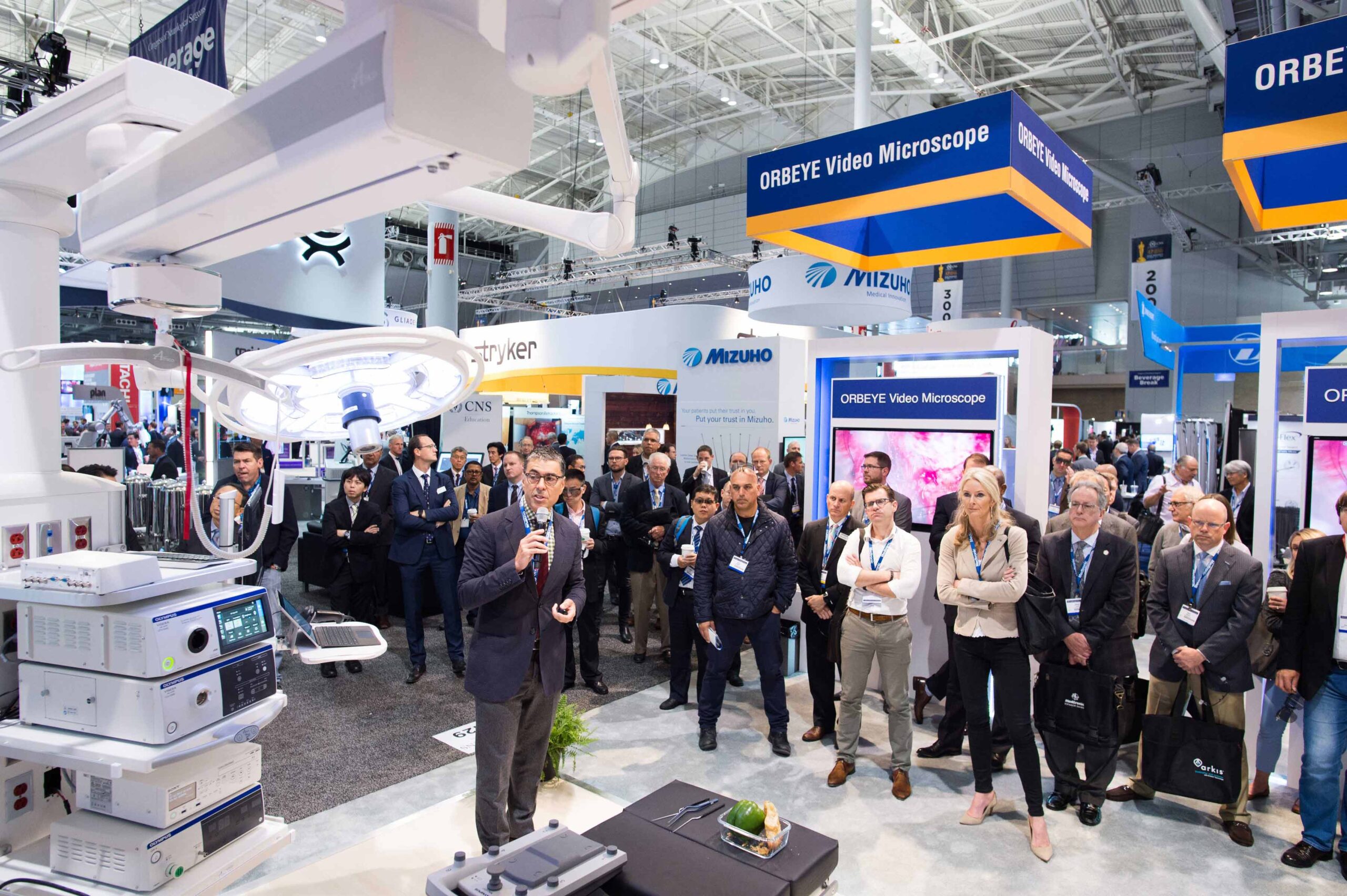 An exhibitor demonstrates a medical product at a trade show on the showroom floor as a large crowd watches.  