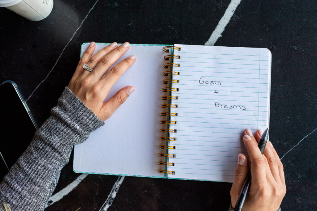 Words "Goals and Dreams" written on a notebook page by a woman holding a black pen