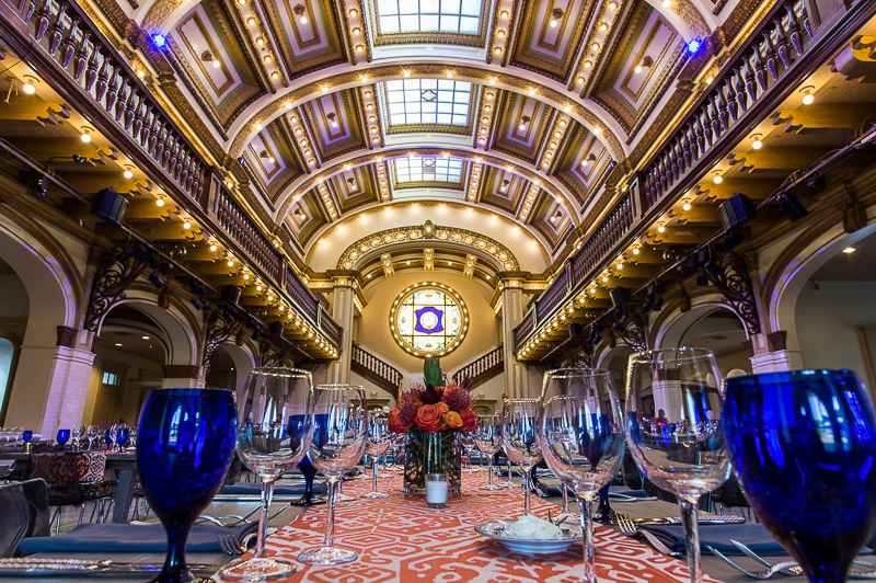 An elegant banquet hall with a grand arched ceiling, intricate architecture, and a long dining table adorned with blue glasses and vibrant floral centerpieces