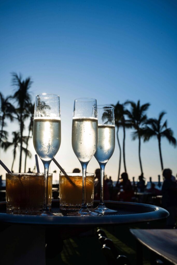 Glasses of champagne and cocktails on a tray with a sunset backdrop and silhouettes of palm trees and people