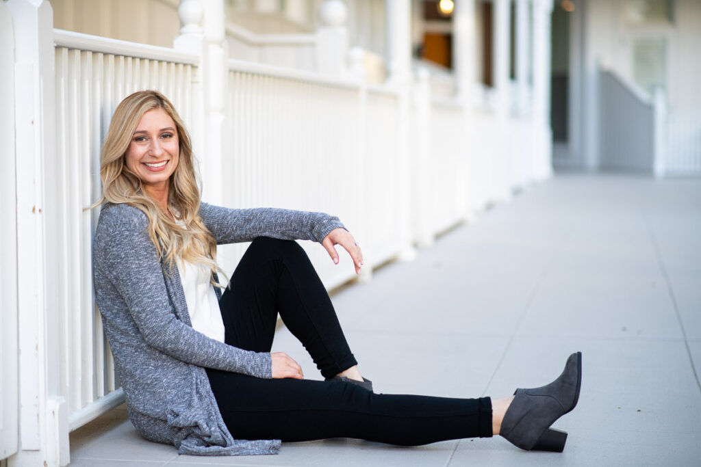 A smiling woman sitting casually on a porch with white railings, wearing a gray cardigan and black pants