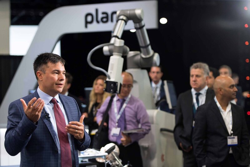 A man in a suit and tie stands beside a robot, explaining to the crowd