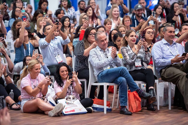 A diverse crowd of individuals seated in chairs, all raising their hands while holding a mobile phone