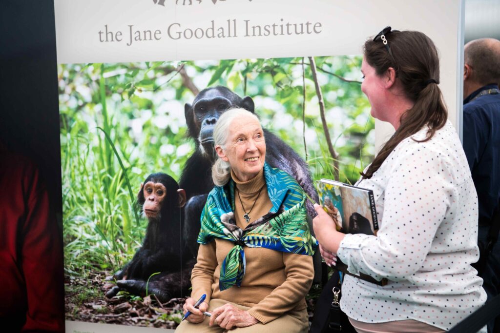 Jane Goodall smiling while engaging with a visitor at an event booth featuring chimpanzee imagery