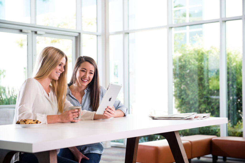 Two women sitting at a table, laughing while looking at a tablet in a bright, modern setting