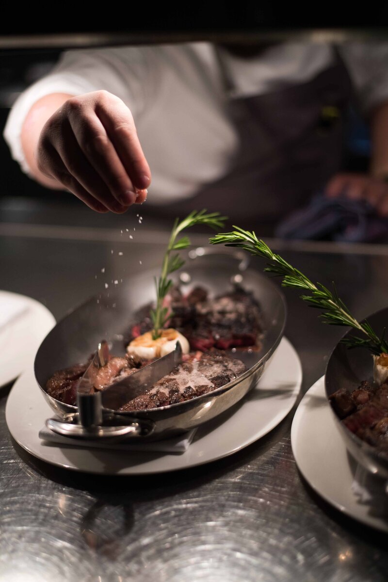 A chef's hand sprinkling salt over a sizzling steak garnished with fresh rosemary