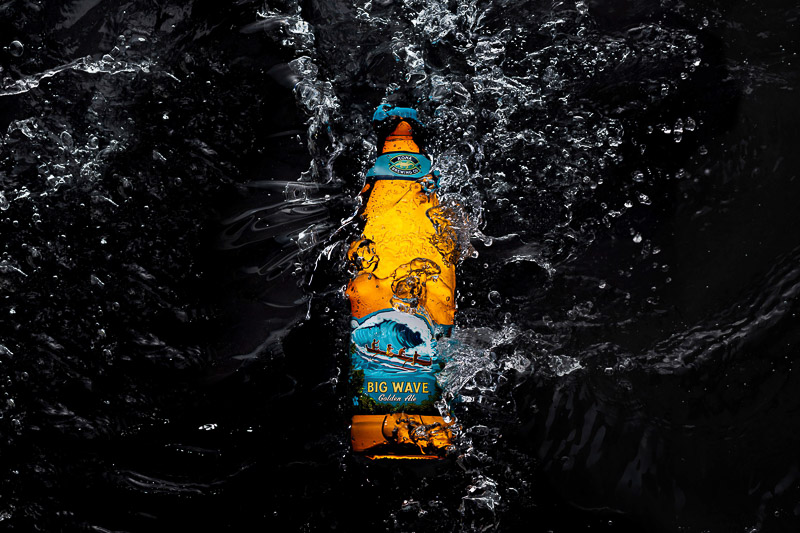 A beer bottle labeled "Big Wave" surrounded by splashing water on a dark background